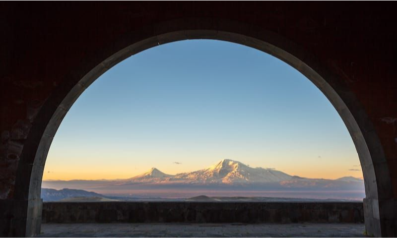 A dramatic view of the snow-capped peaks of Mount Ararat at sunset, framed perfectly through the massive stone archway of the Arch of Charents monument in Armenia during a luxury private heritage tour with Lux Orient.