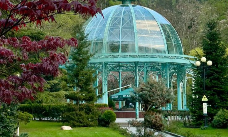 The historic turquoise iron and glass dome mineral spring pavilion surrounded by lush green pine trees in Borjomi Central Park during a luxury private tour in Georgia with Lux Orient.