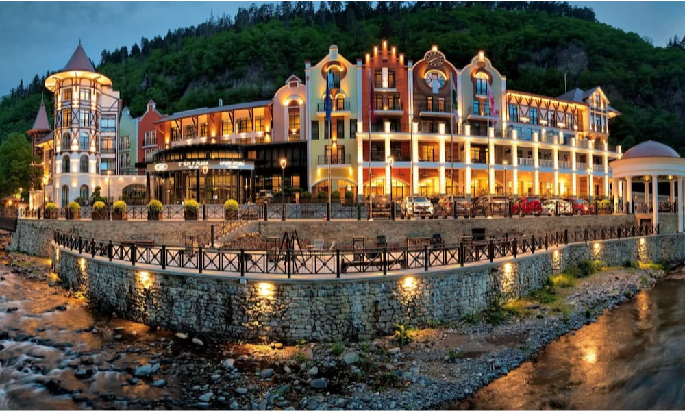 The illuminated fairy-tale architecture of a luxury hotel reflecting on the stone riverbanks of Borjomi town in Georgia at twilight during a bespoke private holiday with Lux Orient.