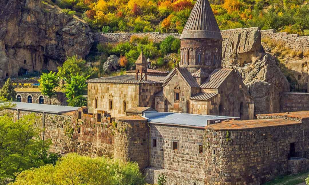 The stone fortress walls, chapels, and prominent conical dome of the medieval Geghard Monastery in Armenia, built against a rugged cliff face with autumn foliage during a luxury private tour with Lux Orient.