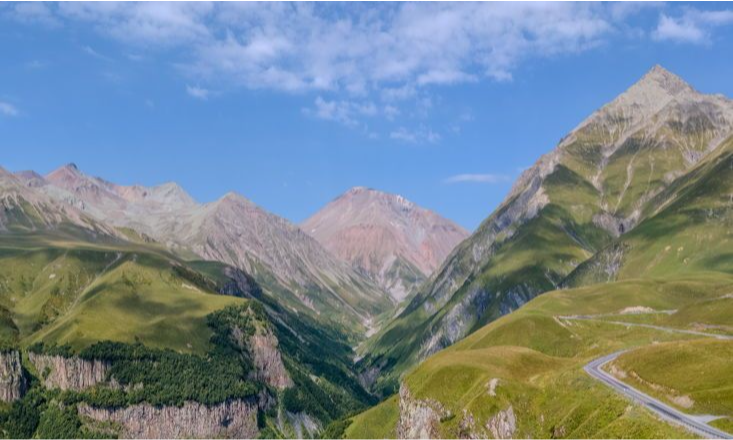 A winding asphalt road twisting along a lush green alpine valley surrounded by dramatic, towering mountain peaks under a blue sky in Gudauri, Georgia during a luxury private tour with Lux Orient.