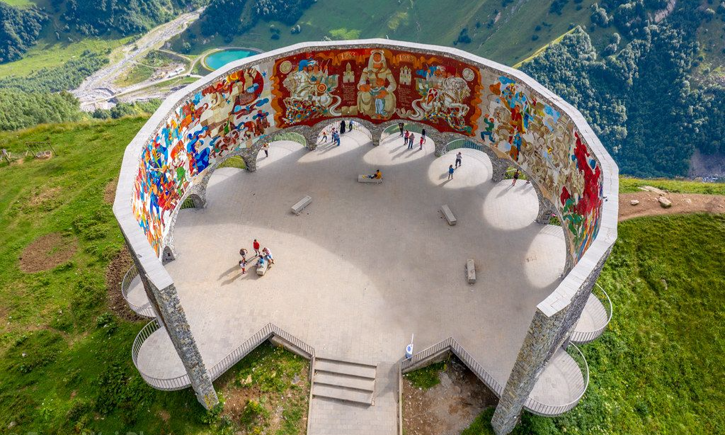 An aerial view looking down at the massive, semi-circular concrete Russia-Georgia Friendship Monument near Kazbegi, displaying its vibrant tiled interior mural on a high mountain cliff during a luxury private tour with Lux Orient.