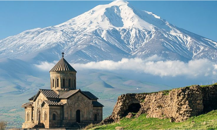 The detailed stone facade and conical dome of Khor Virap Monastery chapel in Armenia standing on a grassy hill near ancient stone ruins under the massive snow-covered peak of Mount Ararat during a luxury private tour with Lux Orient.