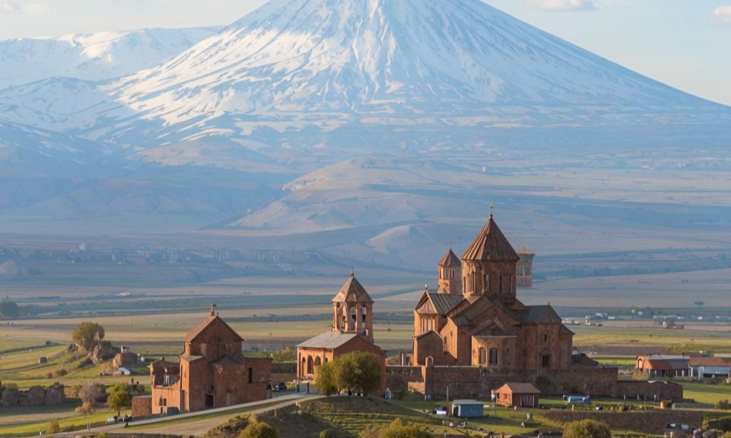 The conical domes and historic brick walls of Khor Virap Monastery complex in Armenia standing on a valley plain beneath the massive snow-capped peak of Mount Ararat during a luxury private tour with Lux Orient.