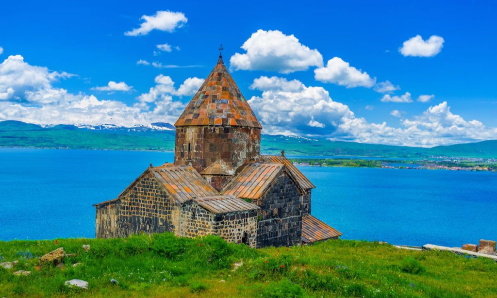 The dark stone brick facade and conical roof of the medieval Sevanavank Monastery in Armenia standing on a green grassy hill overlooking the bright blue waters of Lake Sevan during a luxury private tour with Lux Orient.