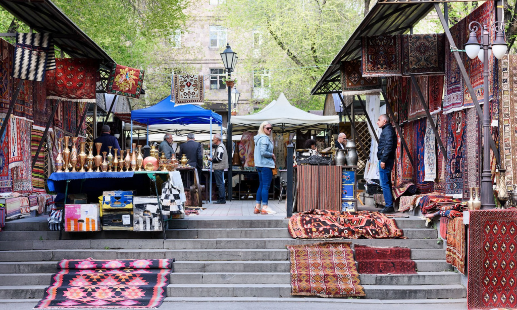 An outdoor market stall at the Vernissage Open Market in Yerevan, Armenia, displaying traditional embroidered hats, vests, and handmade carpets during a luxury private heritage tour with Lux Orient.