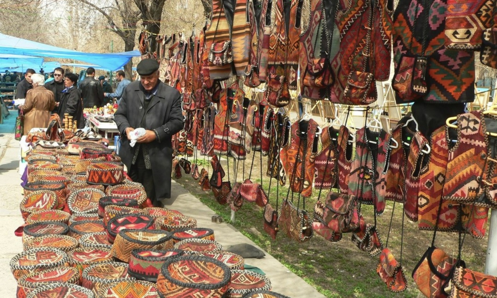 An outdoor market stall at the Vernissage Open Market in Yerevan, Armenia, displaying traditional embroidered hats, vests, and handmade carpets during a luxury private heritage tour with Lux Orient.