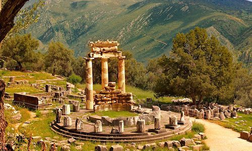 The circular ruins and remaining three Doric columns of the ancient Tholos of Athena Pronaia at the Delphi archaeological site, surrounded by green mountains and olive trees during a luxury bespoke heritage tour with Lux Orient.