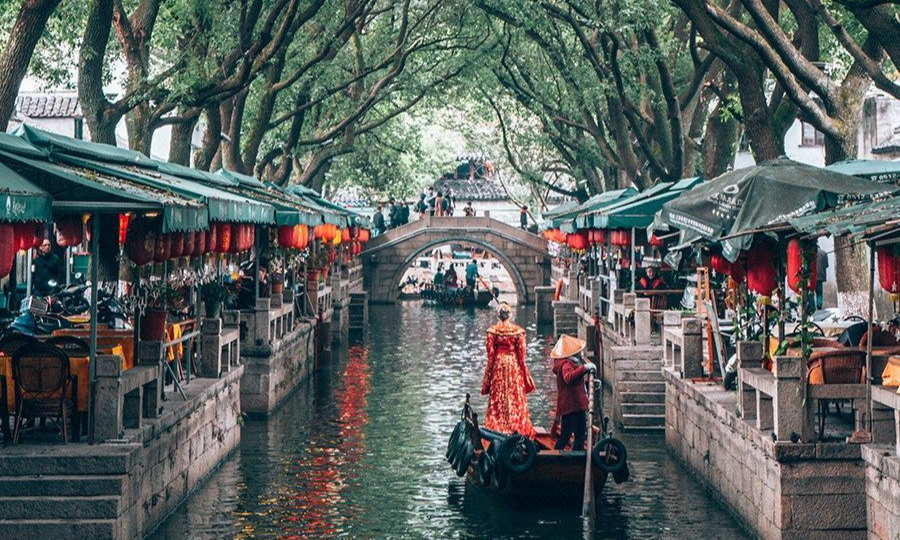 A woman in a traditional red gown stands on a private wooden boat gliding down a tree-lined canal flanked by historic stone buildings and red lanterns in Tongli Water Town during a luxury bespoke tour with Lux Orient.