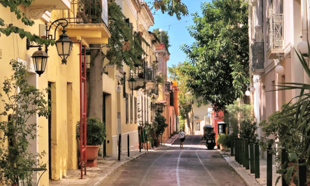 A scenic street scene in the historic Plaka neighborhood of Athens featuring yellow neoclassical buildings shaded by vibrant pink bougainvillea flowers during a luxury private tour with Lux Orient.