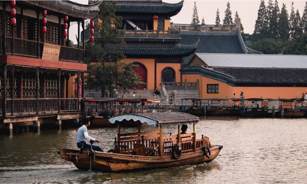 A traditional wooden tourist boat glides under the stone arch of Anlan Bridge, surrounded by historic riverside houses and red lanterns in Zhujiajiao Water Town during a luxury private tour with Lux Orient.