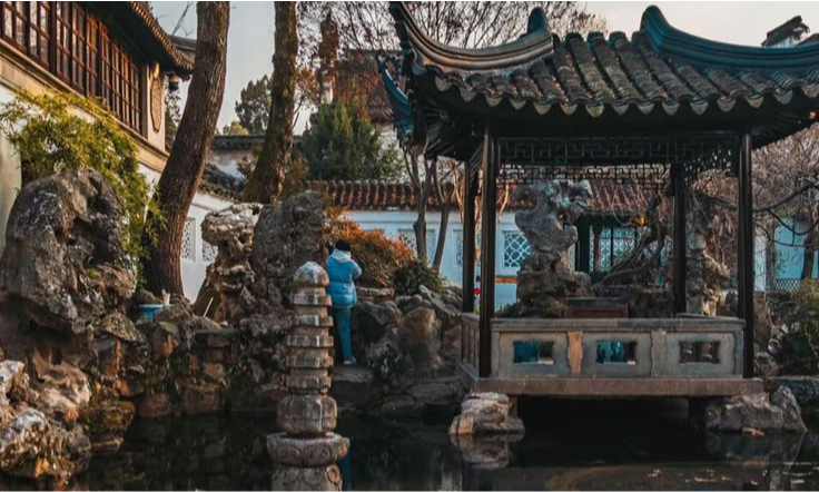 A woman in a traditional red gown stands on a private wooden boat gliding down a tree-lined canal flanked by historic stone buildings and red lanterns in Tongli Water Town during a luxury bespoke tour with Lux Orient.
