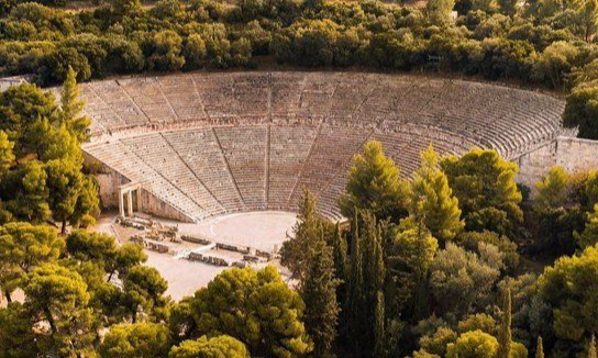 An aerial view of the massive stone semicircular seating tiers of the Ancient Theatre of Epidaurus surrounded by green pine trees during an exclusive luxury private tour from Athens with Lux Orient.
