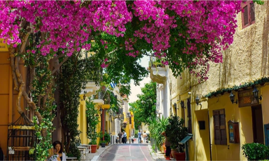 A scenic street scene in the historic Plaka neighborhood of Athens featuring yellow neoclassical buildings shaded by vibrant pink bougainvillea flowers during a luxury private tour with Lux Orient.