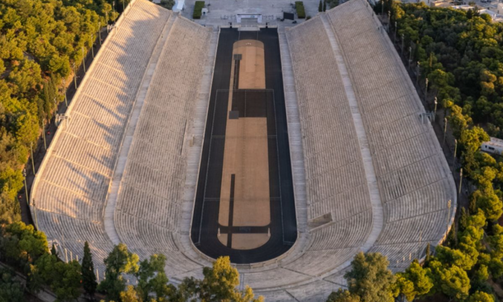 A dramatic aerial view looking down at the long, U-shaped white marble tiers and black running track of the historic Panathenaic Stadium in Athens during a bespoke luxury private tour with Lux Orient.