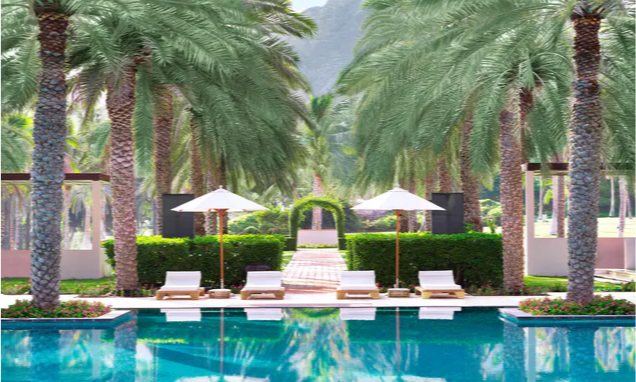 The majestic beachfront exterior and sprawling lagoon pools of Al Bustan Palace, a Ritz-Carlton Hotel, framed by towering palm trees and the rugged Al Hajar Mountains in Muscat, Oman, curated for a VIP itinerary by Lux Orient.
