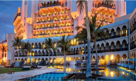The majestic beachfront exterior and sprawling lagoon pools of Al Bustan Palace, a Ritz-Carlton Hotel, framed by towering palm trees and the rugged Al Hajar Mountains in Muscat, Oman, curated for a VIP itinerary by Lux Orient.