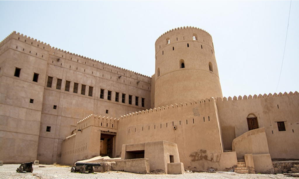 The massive stone facade and towering cylindrical battlements of Al Hazm Castle surrounded by palm groves in Rustaq, Oman, during an elite private luxury heritage tour with Lux Orient