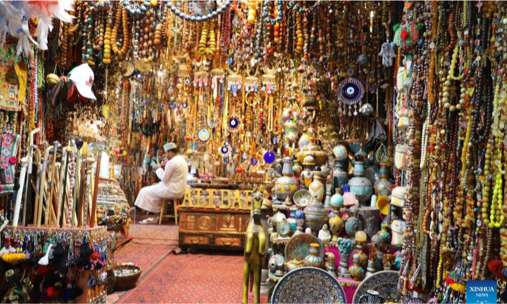The vibrant internal alleyways of Mutrah Souq in Muscat, Oman, showcasing stalls filled with traditional textiles, garments, and hanging bags under a decorative wooden ceiling, arranged by Lux Orient.