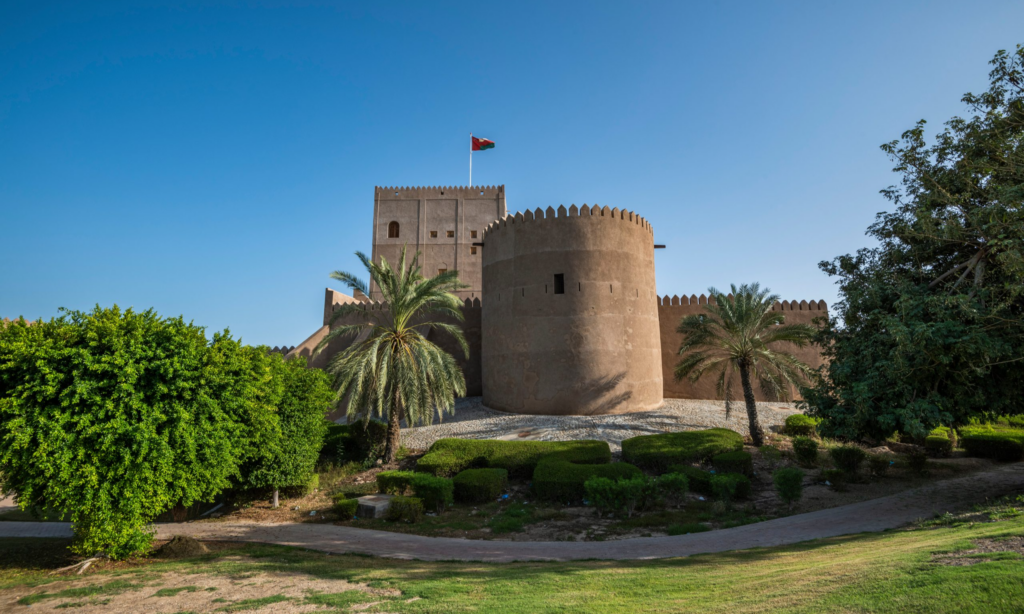 The striking white facade and defensive towers of the historic Sohar Fort surrounded by green palm trees in Sohar Town, Oman, during an exclusive private luxury heritage tour with Lux Orient.