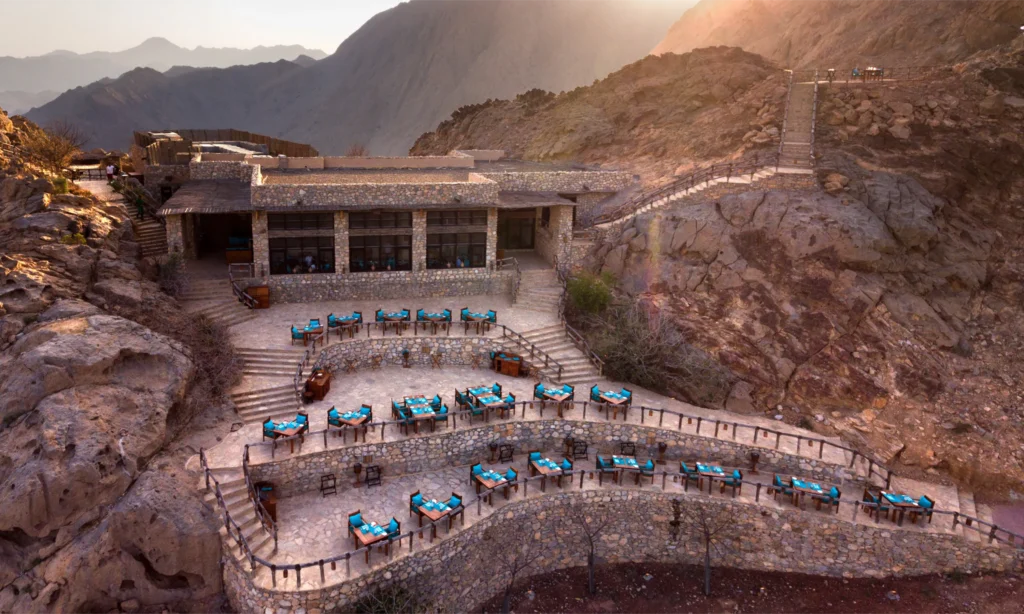 An aerial panoramic view of Six Senses Zighy Bay resort nestled between an expansive white sand beach and the massive, steep desert mountains of Musandam, Oman, arranged by Lux Orient.