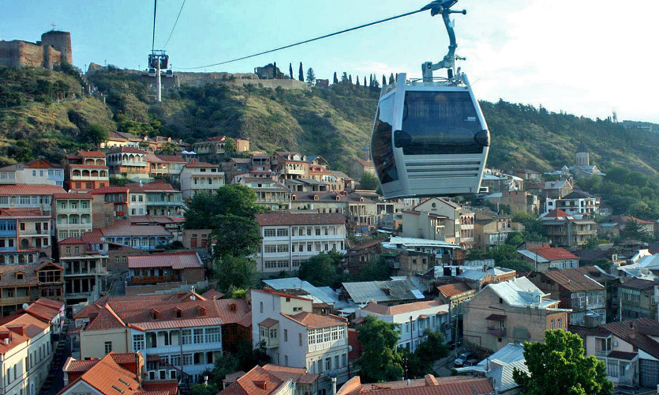 A modern glass aerial cabin of the Tbilisi Cable Car soaring high above the historic brick roofs, winding streets, and scenic riverfront of Old Tbilisi, Georgia, during a luxury private guided city tour with Lux Orient.