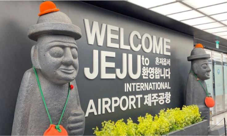 Two traditional Dol Hareubang volcanic stone basalt statues decorated with orange pouches standing next to the welcome sign inside Jeju International Airport in South Korea during a luxury private tour with Lux Orient.