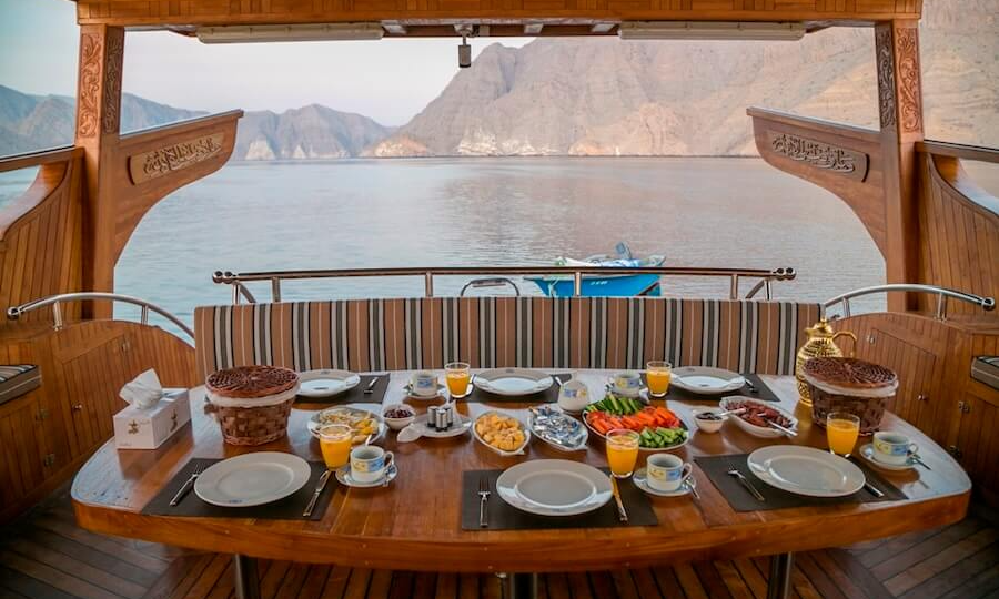 A beautifully dressed luxury dining table laden with gourmet Omani dishes on the open deck of a private Dhow sailing through the scenic fjords of Musandam, Oman, during a VIP charter with Lux Orient.